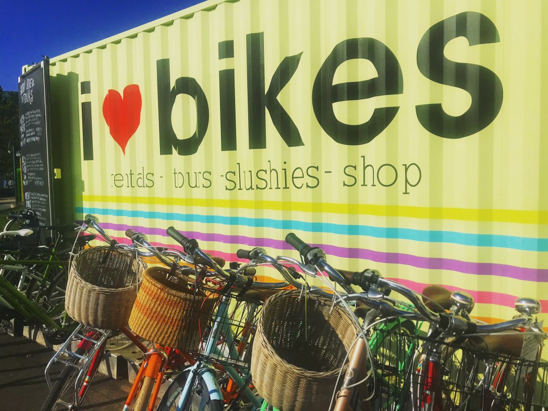 A row of bicycles are lined up against a trailer that reads i heart bikes.