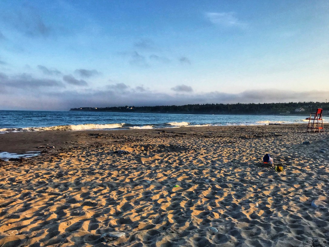 The sun has started to set on a beautiful summer night at Rainbow Haven Beach. Shadows are cast on the brown sand and blue ocean water.